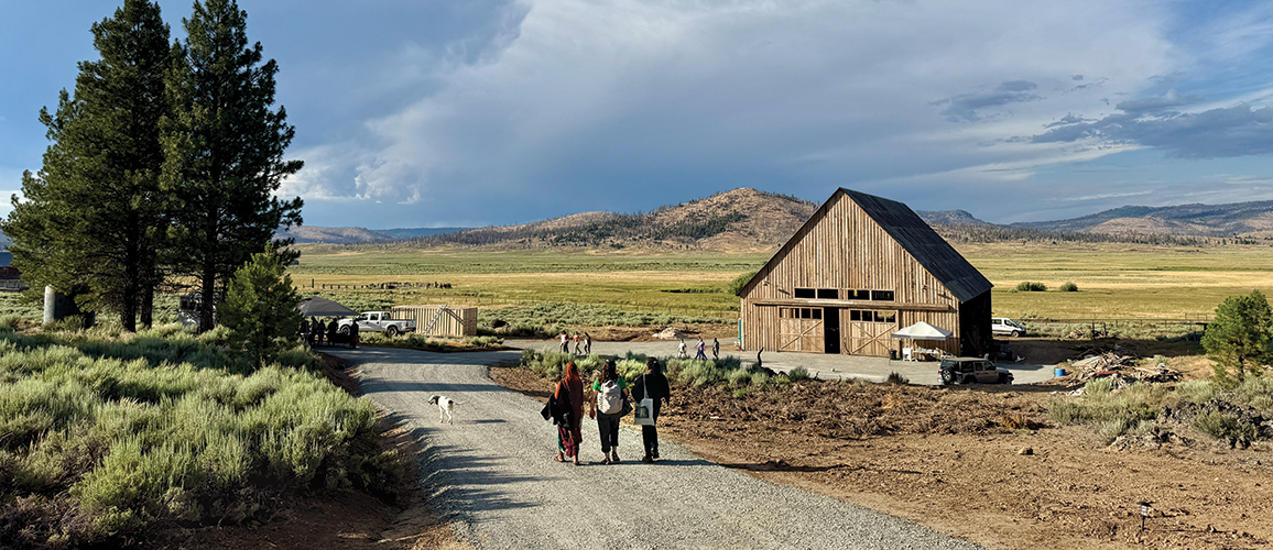 People walking down a gravel road towards a barn illuminated by the evening and backed by clouds.
