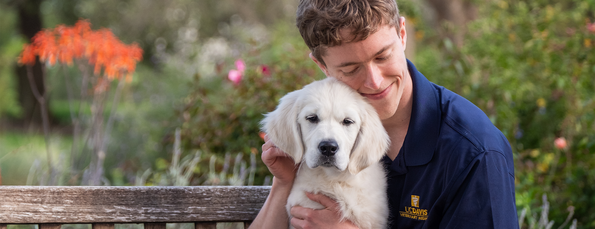 A veterinary student sitting on a bench in the arboretum cuddling a puppy.