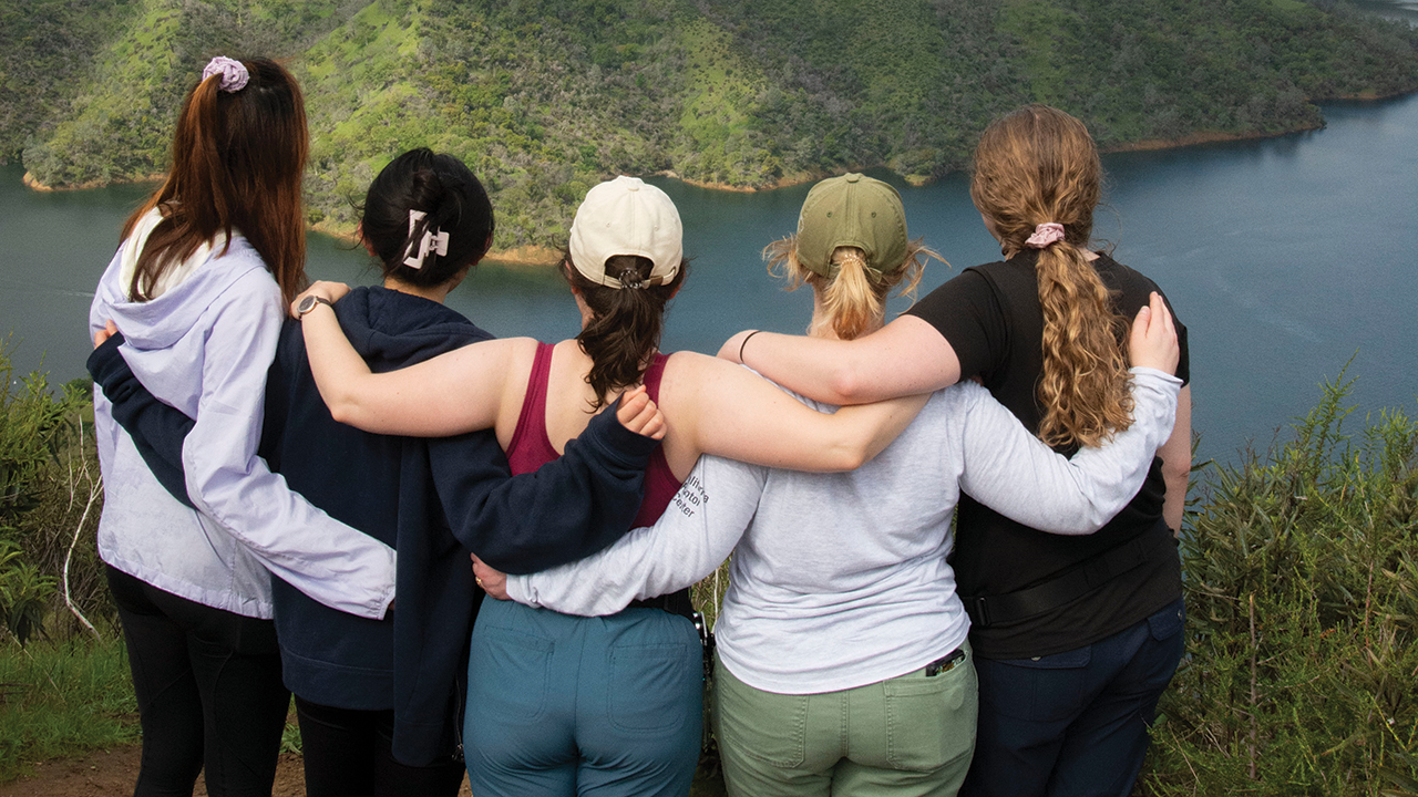 A group of five vet students with arms around each other stand at top of a hill overlooking a lake.