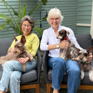 Two smiling older women on an outdoor sofa, each holding a small dog