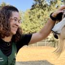 Smiling woman with curly hair and glasses pets a white goat in a sunny farmyard