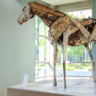 A sculpture of a horse made out of metal standing tall on a platform inside a building, looking out the window.